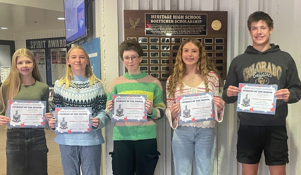 Five high school students stand in a row indoors holding “Heritage High School Student of the Month” certificates. They are smiling and dressed casually in sweaters, jeans, and sneakers, standing in front of a wall display with plaques ."