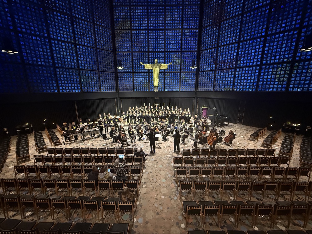 Large student orchestra and choir perform in a modern chapel with blue honeycomb stained-glass walls and a gold statue.