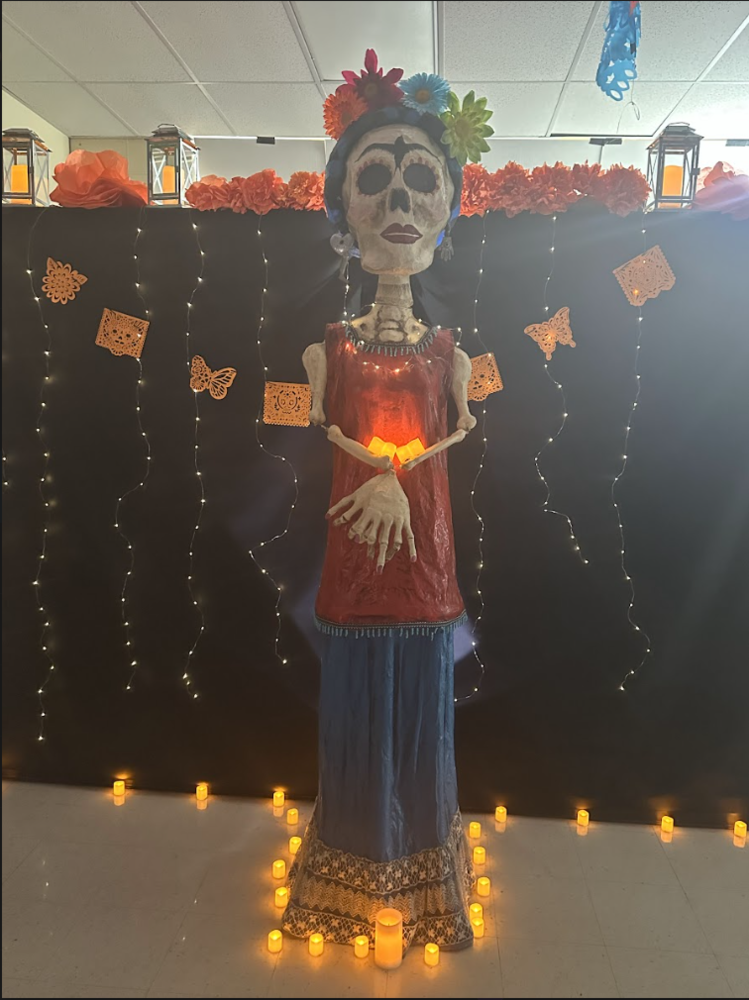 Tall Día de los Muertos Catrina figure dressed in a red blouse and long blue skirt, decorated with lace at the bottom. The skeleton figure has flowers on its head, earrings, and holds a small glowing light at its chest. It stands in front of a black backdrop with string lights, orange paper marigolds, papel picado banners, lanterns, and surrounded by small electric candles on the floor.