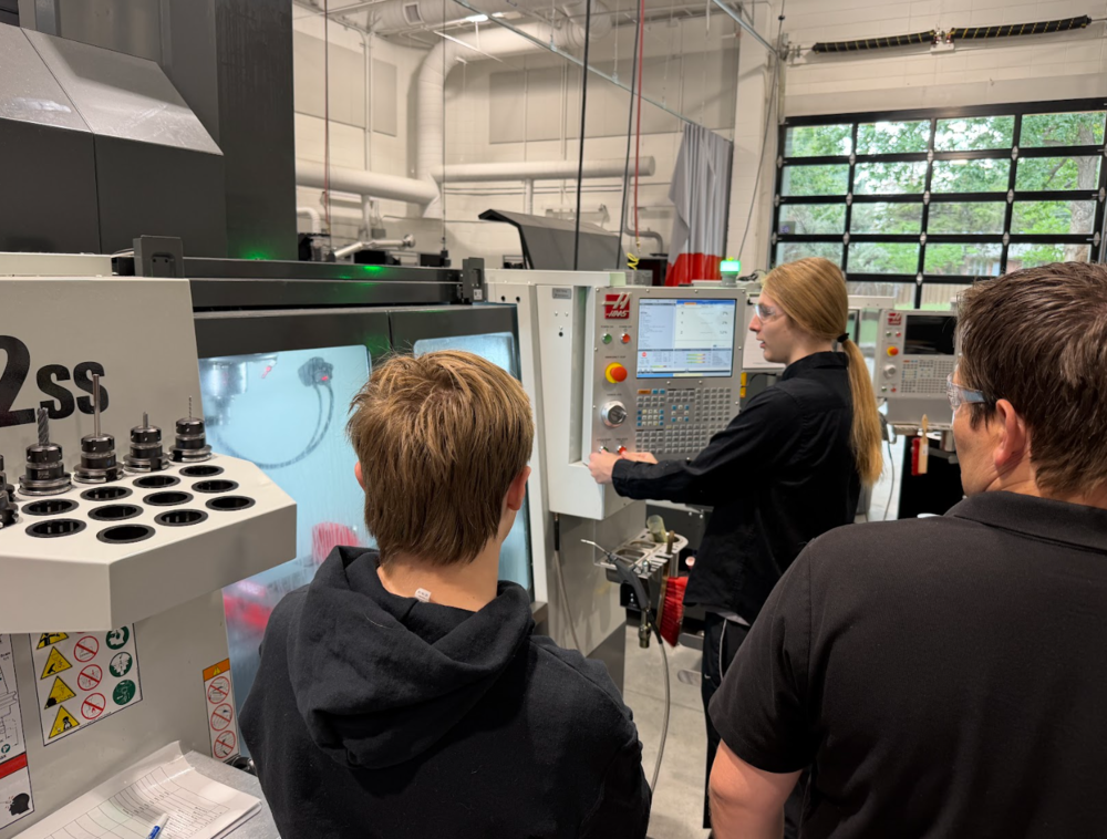 A student wearing safety glasses operates a large Haas CNC machine control panel while two individuals observe.
