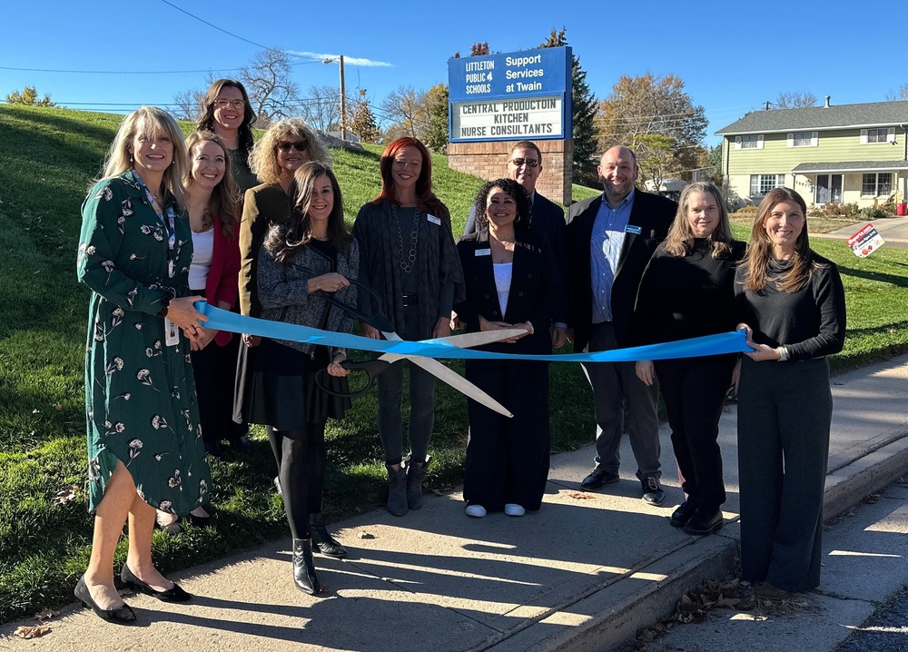A group of eleven people cutting a large blue ribbon in front of a blue business sign on a sunny day.