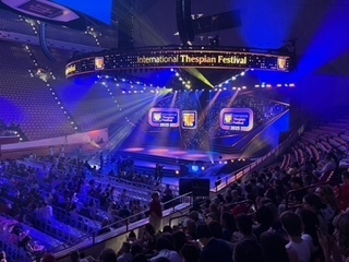 Large indoor arena with audience seating and a lit stage reading “International Thespian Festival” with event screens.