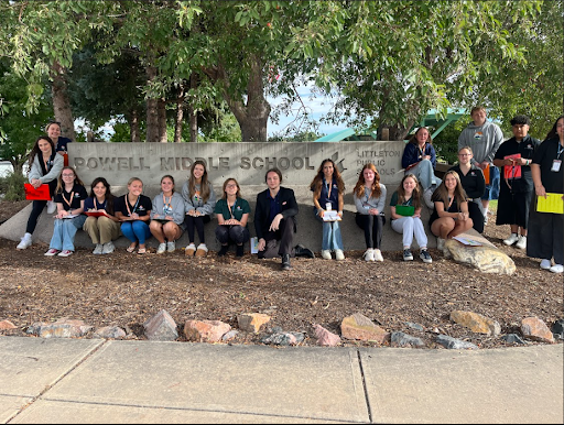  A group of about fifteen high school students and a teacher are posing for a picture in front of a stone sign for Powell Middle School. Many of the students are crouched down in a row in front of the sign.