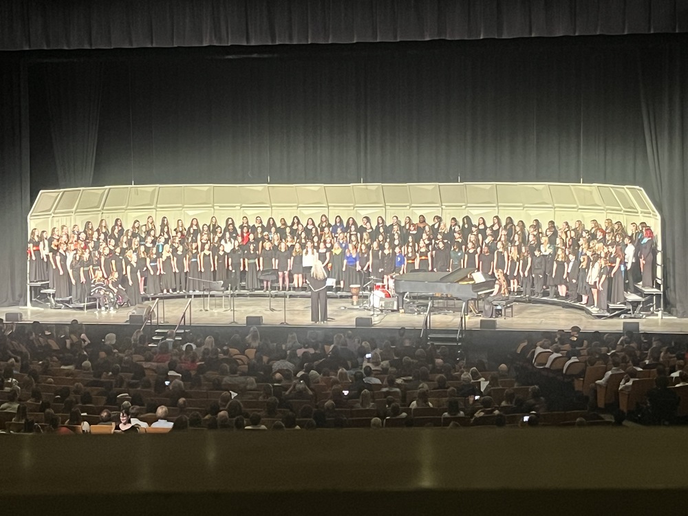 A large student choir performs on a stage with a conductor and grand piano before a seated audience in a theater.
