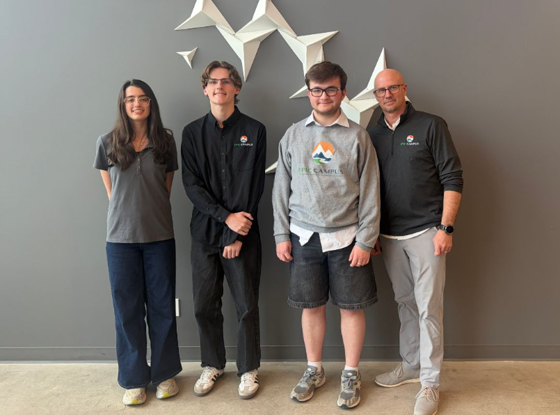 Three students and one adult standing in front of a gray wall with white geometric art.