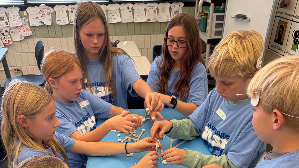 Group of seven elementary-aged students wearing matching blue “Heritage HSS” shirts gather around a table covered in a blue tablecloth. They are building structures using marshmallows and wooden sticks in a classroom setting. The students are focused and engaged, collaborating on the activity. In the background, decorated T-shirts hang along the wall under a window.