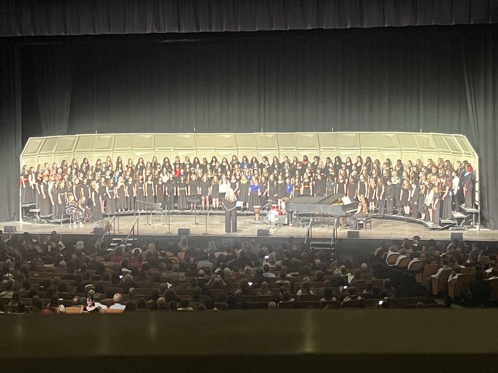 A large student choir performs on a stage with a conductor and grand piano before a seated audience in a theater.