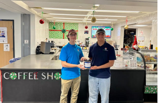 Two students stand in a decorated coffee shop, one holding a jar and the other accepting it.