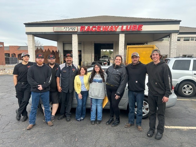 A group of seven adults and one teenager posing for a photo in front of the Raceway Lube auto shop.
