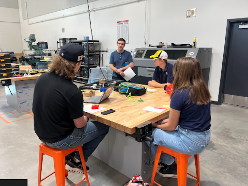 Three students sit at a workbench with laptops and materials, facing an instructor who stands behind the table in a workshop setting.