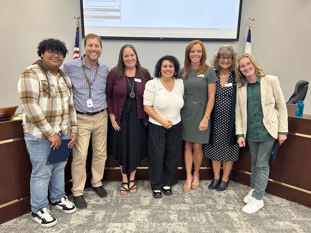 Two students stand at each end of a group photo with the Board of Education; diverse in age, gender, and ethnicity; flags and screen in background.