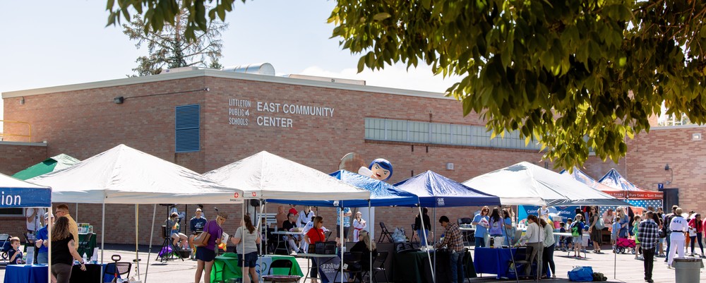 Community members visit outdoor booths set up under white and blue tents at the East Community Center during a sunny day. The brick building with the sign 'Littleton Public Schools East Community Center' is visible in the background, and people are engaging with local organizations and resources.