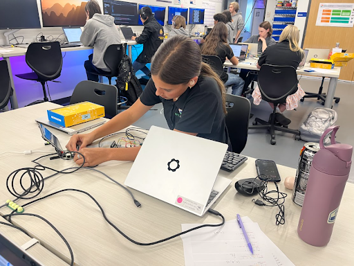 A close-up of a female student working with a small electronic device connected to wires and a laptop on a cluttered desk in a busy computer lab.