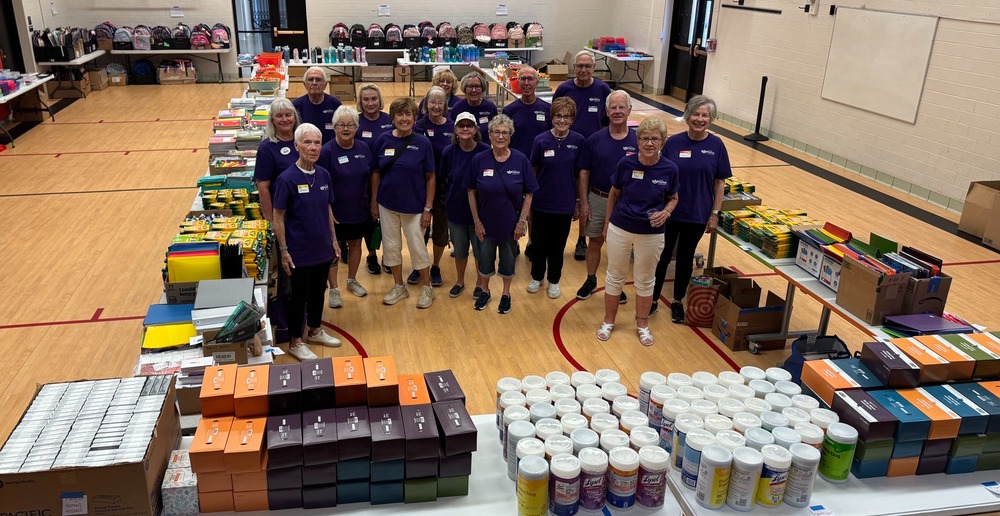 Volunteers in matching purple shirts pose in a gymnasium surrounded by organized school supplies and backpacks.