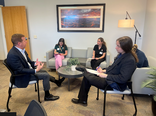 A man in a suit speaks to two people across a coffee table while two young women listen from a nearby couch in a casual office reception area.