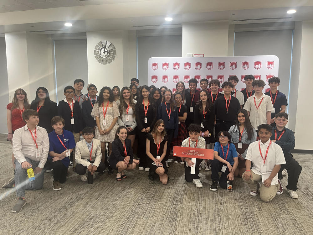 A group of diverse middle school students from Euclid Middle School posing indoors at a Latinos in Action conference. Most students wear semi-formal attire with red lanyards and ID badges. A red sign reading "Euclid Middle School" is held at the front. Behind the group is a branded backdrop with a repeated red and white logo featuring silhouetted figures. The setting is a modern conference room with neutral walls, carpeting, and a clock on the wall.