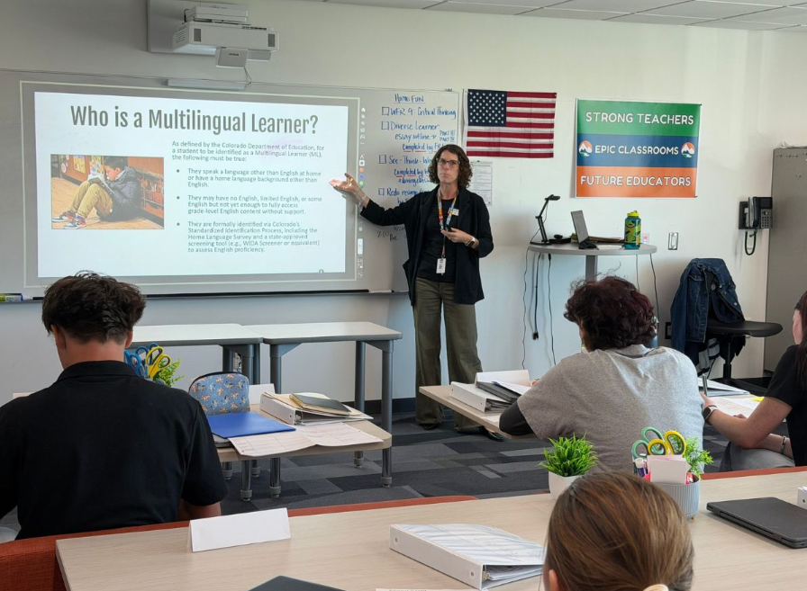 Teacher presents a slide titled "Who is a Multilingual Learner?" to a classroom of students seated at desks.