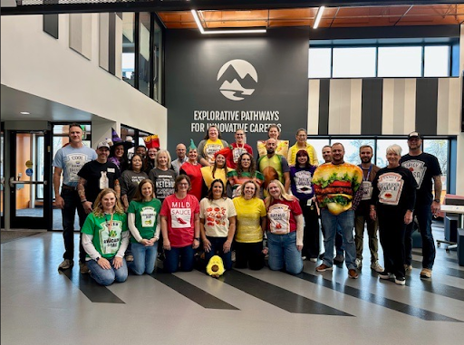 A large group of people in food-themed Halloween costumes posing for a photo in a modern office lobby with the "Explorative Pathways for Innovative Careers" logo on the wall.