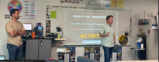 Two men stand in a classroom, one presenting a slide titled "Reality vs. Perception" on a projection screen.