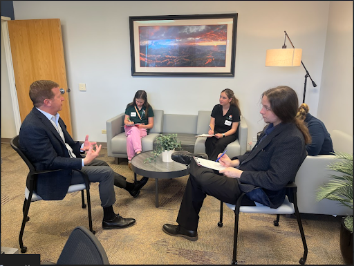 A man in a suit speaks to two people across a coffee table while two young women listen from a nearby couch in a casual office reception area.