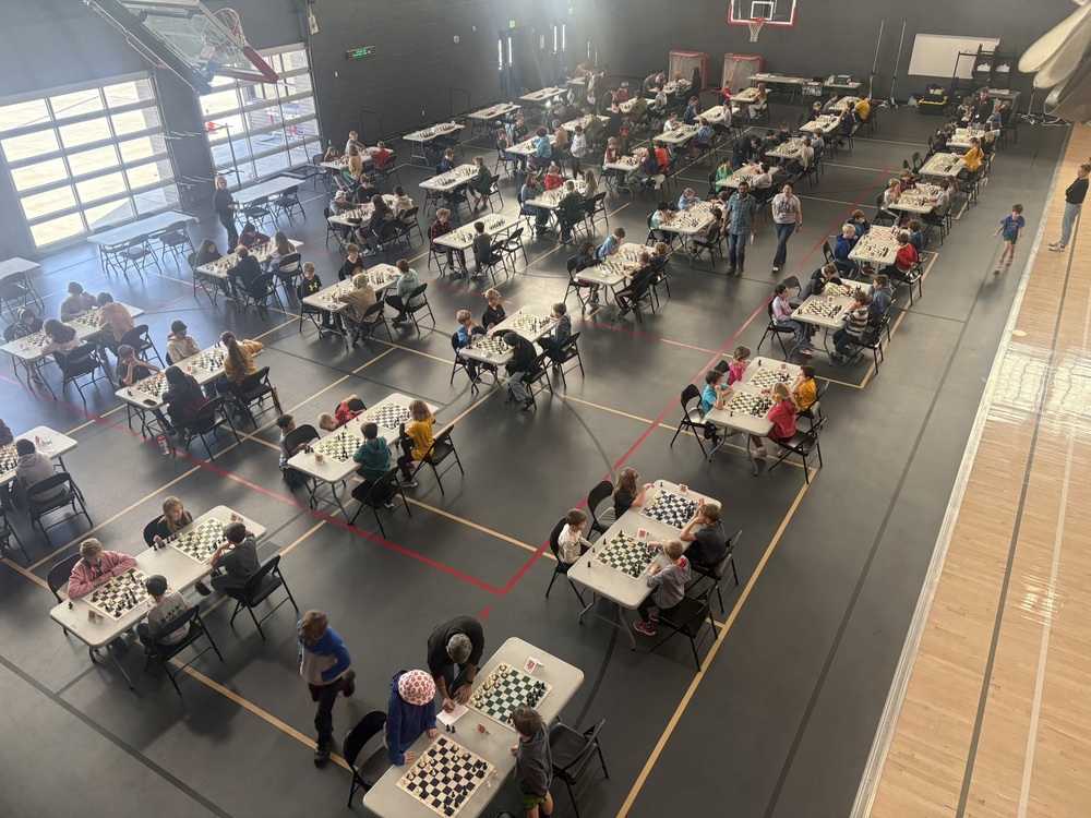 A large group of students sit at small tables playing chess during a tournament in a school gymnasium.