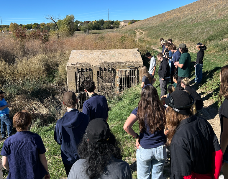 Students stand outdoors on a grassy hillside, looking down at a concrete water structure with metal grates.