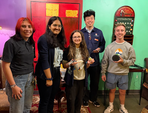 Five students stand together in a colorful room, posing with a small electronic project and a black speaker.