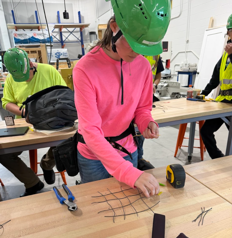 Student in a pink shirt and green hard hat using wire to lay out and twine together a micro scale for rebar.