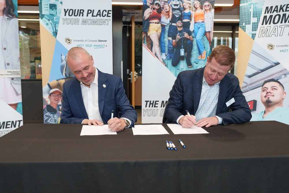 Two men in suits sit at a black table, smiling as they sign official partnership documents with pens.
