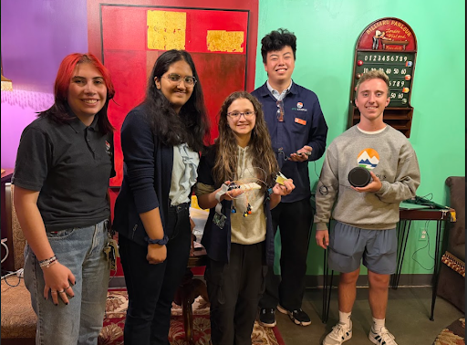 Five students stand together in a colorful room, posing with a small electronic project and a black speaker.
