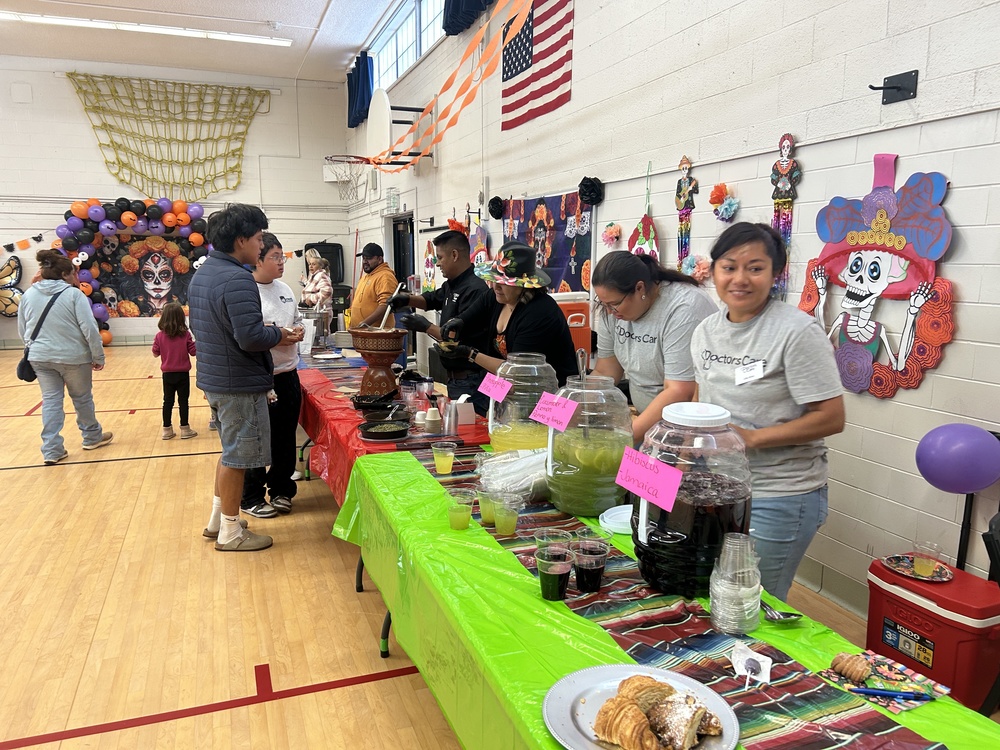 Community event with people at a long table with drinks and food, decorated for Día de Muertos in a gym.
