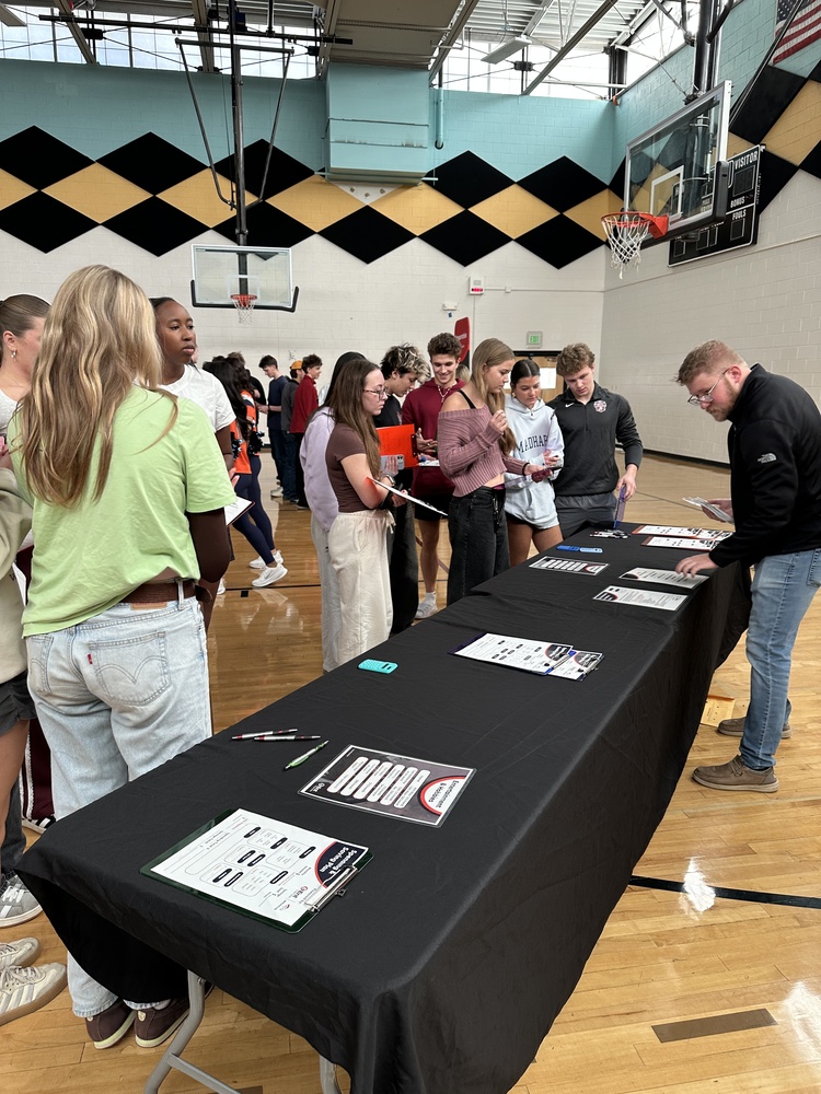 A group of high school students talking with an adult man standing behind a long, black table in a gymnasium.