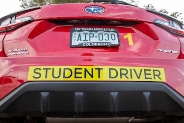 Rear view of a red Subaru Crosstrek with a large yellow “STUDENT DRIVER” sign on the bumper. The car has all-wheel drive branding and a number “1” decal on the back. Trees and cloudy sky are reflected in the vehicle’s surface.