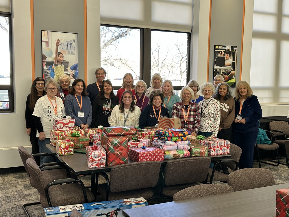 A large group of smiling volunteers stands behind a table stacked high with colorful wrapped holiday gifts and blankets.