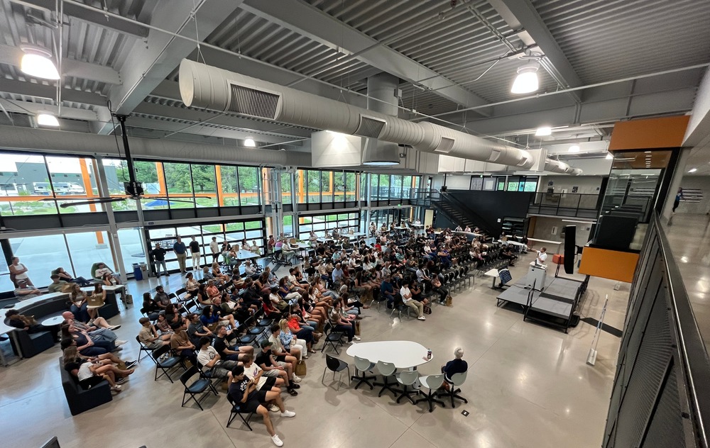 A large group of students and adults sit in a modern, open-concept school commons area with high ceilings and exposed ductwork. Rows of chairs and soft seating fill the space as attendees face a small stage where a speaker stands. Natural light streams through tall windows and glass garage doors, with views of the outdoors visible. Some people stand along the walls or sit at round tables, indicating a school meeting or orientation event.