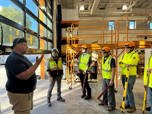 An instructor talks to a group of students wearing safety vests and hard hats in a large indoor construction lab space with scaffolding.