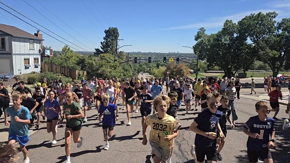 A large group of youth runners competes in a road race running down a suburban street on a sunny day.