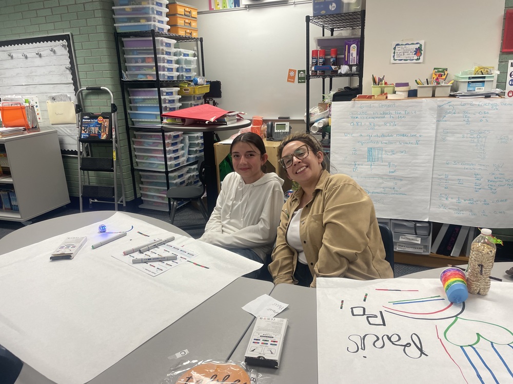 A student and a parent sit side by side at a table in a colorful, well-stocked classroom. The student on the left wears a white hoodie and has long, dark hair tied back. The parent on the right wears glasses, a tan jacket, and a white shirt, smiling at the camera. On the table are large sheets of white paper with colorful marker drawings, robotics-related materials including Ozobot markers and a small robot, and a container of cereal. Behind them, shelves are filled with neatly stacked plastic bins, art supplies, and instructional posters, including a large chart paper with handwritten math notes. The classroom appears inviting and organized.