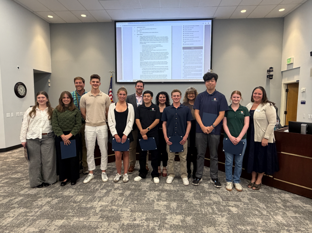 A group of students and adults stand together holding awards in front of a projection screen at an LPS Board meeting.