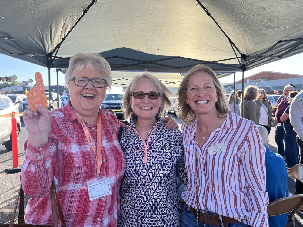 Three adult women smile under a tent at a tailgate party. The woman on the left holds an orange foam finger.