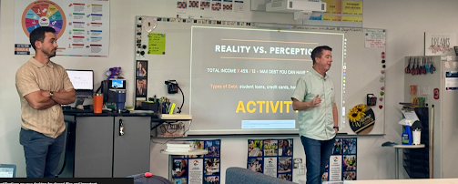 Two men stand in a classroom, one presenting a slide titled "Reality vs. Perception" on a projection screen.