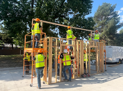 A group of people wearing safety vests and hard hats work together to build a wooden frame structure outdoors, using ladders and scaffolding.