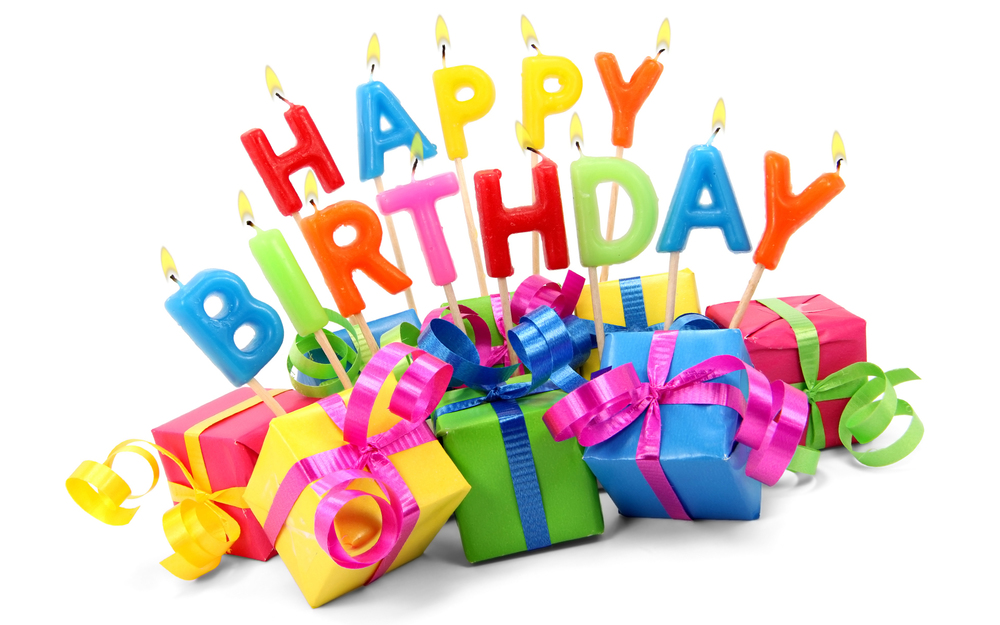 Colorful candles spelling "HAPPY BIRTHDAY" on sticks, each letter in a different bright color. The candles are lit, and the letters are placed above brightly wrapped presents in pink, yellow, green, and blue, each topped with curly ribbons.