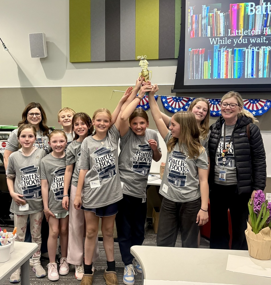 A team of 8 elementary students holds up a Battle of the Books trophy. They are flanked by their two coaches.