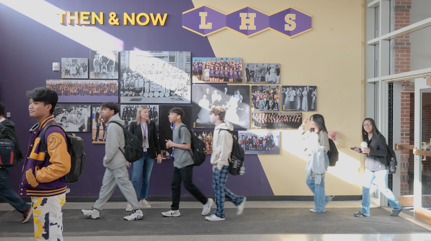 Students walk past a “Then & Now” photo wall at the entrance of Littleton High School, under a purple and gold LHS sign.