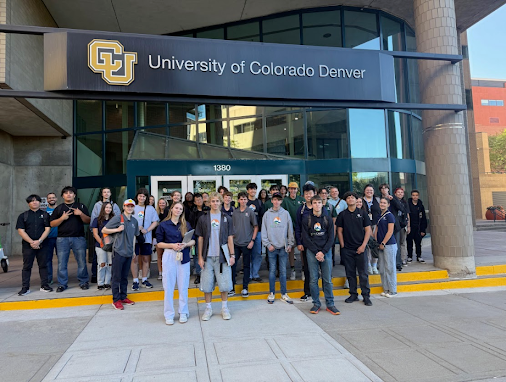  A group of students and adults stand outside a building with a large sign that reads University of Colorado Denver above the entrance. They are posed for a group photo.