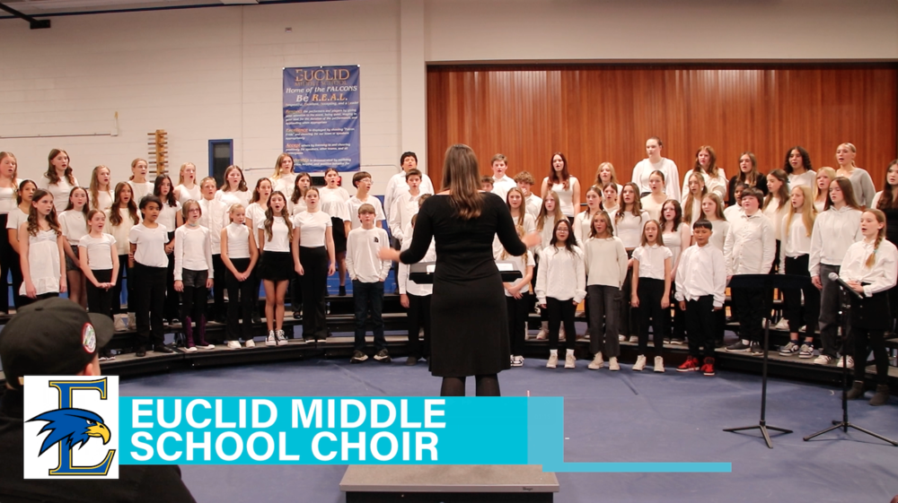 Euclid Middle School choir performing in a school gym or music room. A large group of diverse students stand on tiered risers, all dressed in black and white, facing a conductor with long hair wearing a black dress. A blue banner on the wall behind reads “Euclid Middle School – Home of the Falcons – Be R.E.A.L.” A blue and wood-paneled background frames the scene. A turquoise text banner at the bottom left reads “Euclid Middle School Choir” alongside the school's falcon logo. A microphone stand is visible on the right.