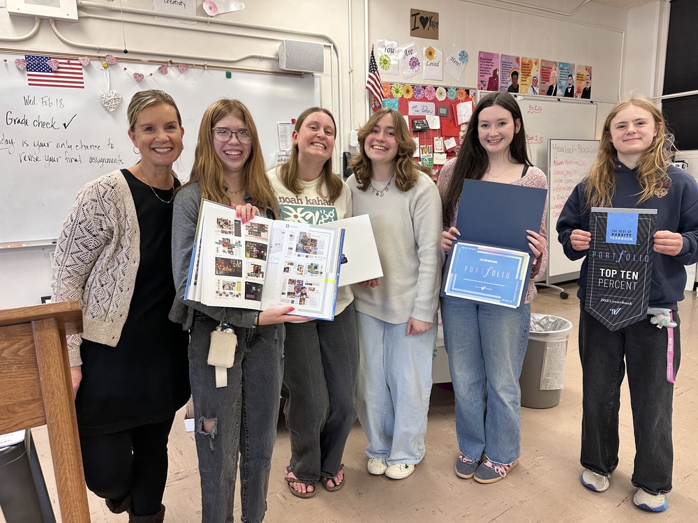 Six smiling students and a teacher posing with a yearbook award and banner in a classroom.