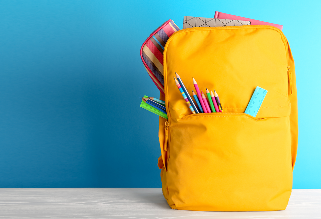 Yellow backpack on a table with colorful pencils, rulers, notebooks, and a striped pencil case against a blue background.
