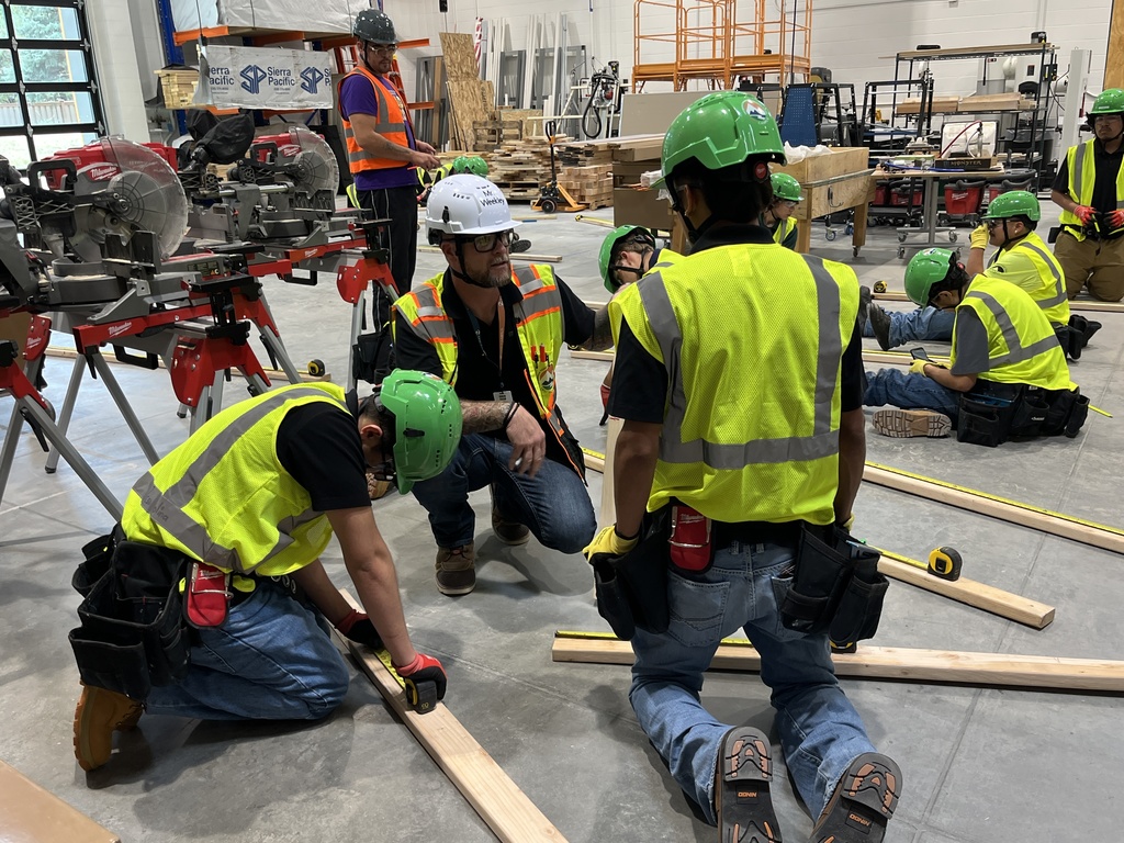 A group of students in a construction workshop wearing green hard hats, yellow reflective vests, jeans, and work boots. They are working in pairs, measuring and aligning wooden planks on the floor. An adult instructor in a white hard hat and reflective vest is kneeling and supervising their work. The workshop contains saws, power tools, and lumber in the background, with additional students working in the back. Safety gear and construction equipment are visible throughout the space.